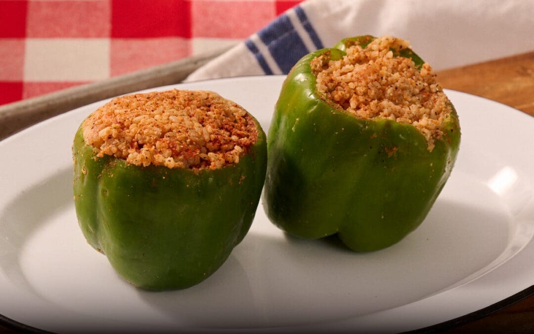 Two seafood-stuffed bell peppers on a white plate, generously seasoned and filled with a flavorful mixture of rice and seafood. A red and white checkered tablecloth and a blue-striped napkin add a cozy, Cajun-inspired touch in the background.